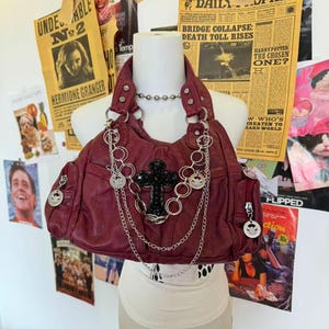 以下が含まれることがあります： シルバーのチェーンとリングのアクセント、黒い十字架、シルバーのコインチャームが付いた、バーガンディ色のレザーハンドバッグ。バッグには2つのジッパー付きサイドポケットがあり、マネキンに展示されています。背景にはヴィンテージの新聞の切り抜きがあります。
