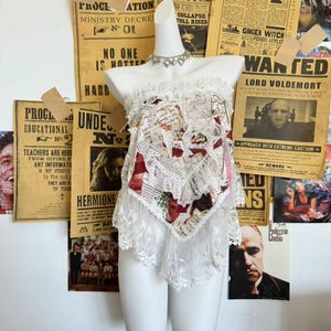 May include: A white mannequin displaying a two-piece outfit. The top is a patchwork of printed fabric with red accents, trimmed with white lace. The bottom is a matching white lace. A silver necklace adorns the mannequin's neck.