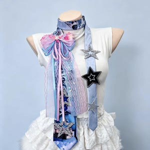 May include: A light blue necktie featuring a large decorative bow made of pink and blue ribbons, lace, and tulle. The tie is adorned with multiple star-shaped patches in silver, black, and glittery finishes, worn over a white sleeveless top.