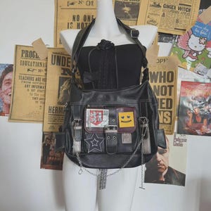 Könnte beinhalten: Schwarze Kunstleder-Umhängetasche mit mehreren Taschen und silbernen Kettenakzenten. Mit Aufnähern mit einem Stern, einem Smiley und einer Karte im Vintage-Stil. Die Tasche wird auf einer Schaufensterpuppe vor Vintage-Zeitungsartikeln präsentiert.