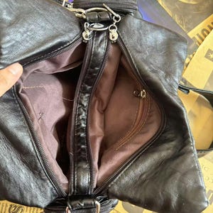 May include: Black leather handbag with multiple compartments and silver-tone hardware. The bag features a zippered main compartment and a smaller zippered pocket. The interior lining is a brown fabric. The bag has a shoulder strap and a small handle.