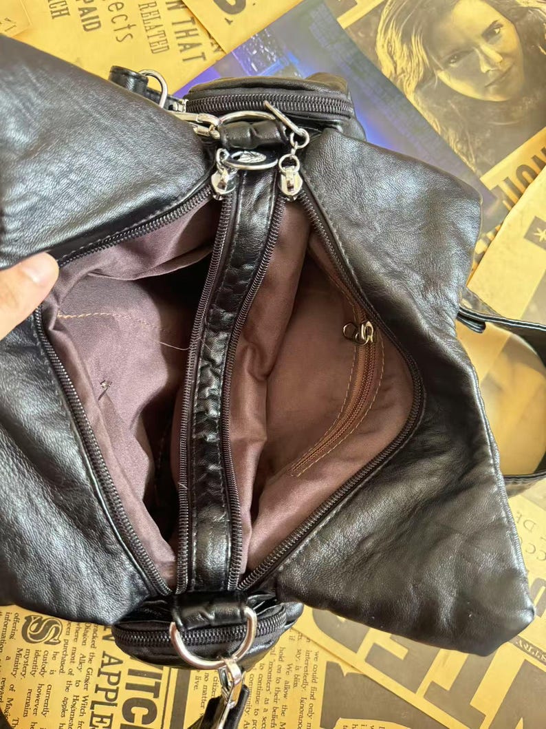 May include: Black leather handbag with multiple compartments and silver-tone hardware. The bag features a zippered main compartment and a smaller zippered pocket. The interior lining is a brown fabric. The bag has a shoulder strap and a small handle.