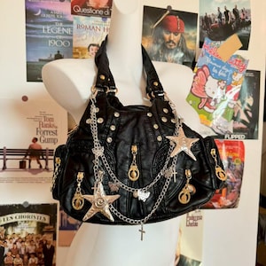 May include: Black faux leather handbag with silver-tone star and butterfly charms, studs, and zippers. The bag has a shoulder strap and multiple pockets. Movie posters are in the background.