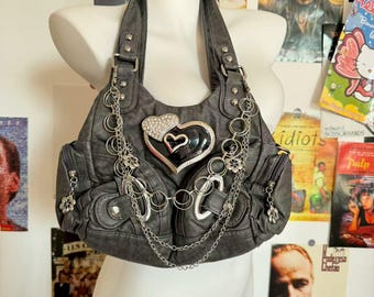 Könnte beinhalten: Graue Handtasche mit silbernen Ketten und Herzdetails. Die Tasche hat eine große herzförmige Verzierung mit Strasssteinen und ein kleineres schwarzes Herz. Sie hat einen Schulterriemen und mehrere Taschen.