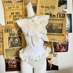 Puede incluir: Un torso de maniquí blanco con una flor decorativa y ribete con volantes. Un collar plateado adorna el cuello. El fondo presenta carteles y recortes de periódicos de estilo vintage, incluidos carteles de "Se busca" y carteles de películas.