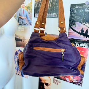 May include: A purple and brown shoulder bag with a gold zipper and brown handles. The bag has a rounded shape and appears to be made of leather. The background includes movie posters.