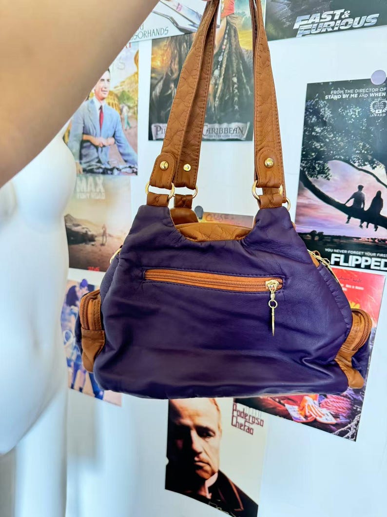 May include: A purple and brown shoulder bag with a gold zipper and brown handles. The bag has a rounded shape and appears to be made of leather. The background includes movie posters.