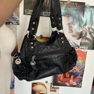 May include: Black handbag with double handles and silver-tone hardware. The bag has a zippered main compartment and multiple exterior pockets. The bag is hanging in front of movie posters.