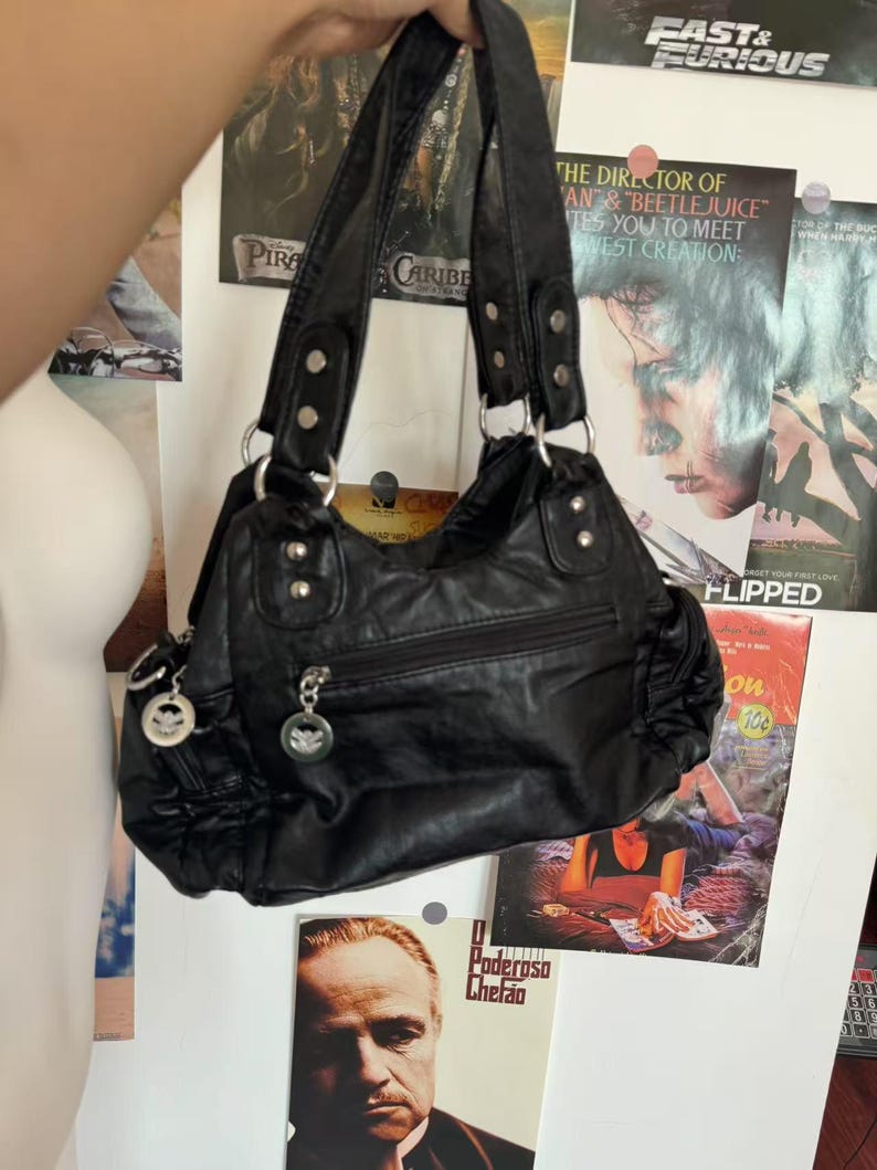 May include: Black handbag with double handles and silver-tone hardware. The bag has a zippered main compartment and multiple exterior pockets. The bag is hanging in front of movie posters.