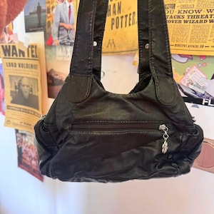 May include: Black leather shoulder bag with a zipped front pocket and a decorative charm. The bag has two shoulder straps and is hanging against a wall with posters.