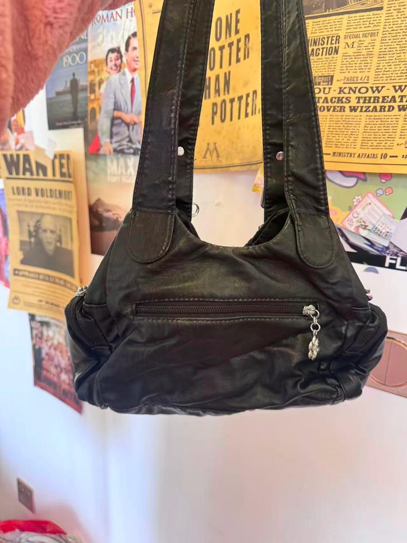 May include: Black leather shoulder bag with a zipped front pocket and a decorative charm. The bag has two shoulder straps and is hanging against a wall with posters.