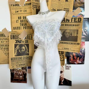 Puede incluir: Un busto blanco exhibe un delicado top de encaje con un escote de corazón y una larga corbata fluida. La parte superior está adornada con encaje intrincado y adornos sutiles. Un collar plateado en forma de corazón añade un toque de elegancia. El fondo presenta carteles vintage.