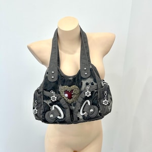 May include: A dark gray handbag with a textured animal print pattern, featuring a prominent central heart-shaped emblem with a deep red center, silver-toned metal hardware, decorative flower-shaped charms, and two sturdy shoulder straps.