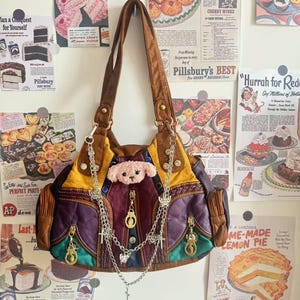 May include: A patchwork shoulder bag in shades of brown, yellow, purple, and green, adorned with a teddy bear and chains. The bag has gold-toned zips and hardware. Vintage advertisements form the backdrop.