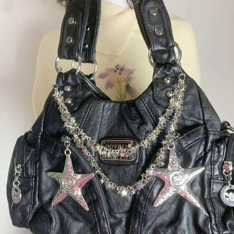 May include: Black leather handbag with silver star charms and a chain of small stars. The bag has two handles, a zipper pocket, and a decorative label that reads "OUYA".