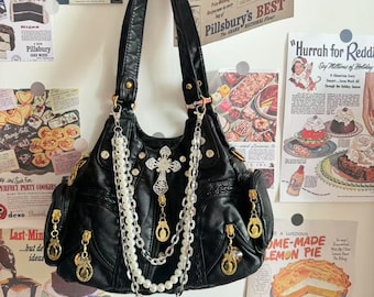 Könnte beinhalten: Schwarze Handtasche aus Kunstleder mit goldfarbener Hardware, einem Kreuzanhänger und perlenartigen Kettenakzenten. Die Tasche hat mehrere Taschen und einen oberen Griff. Vintage-Anzeigen sind im Hintergrund.