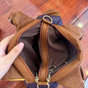 May include: A brown leather handbag with a purple accent. The bag has multiple zippered compartments and gold-colored hardware. The interior is a dark brown color.