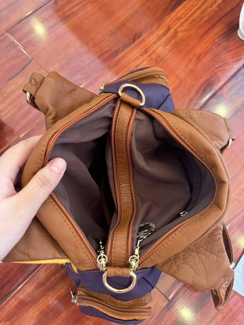 May include: A brown leather handbag with a purple accent. The bag has multiple zippered compartments and gold-colored hardware. The interior is a dark brown color.