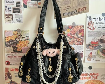 May include: Black leather handbag with gold-tone hardware, featuring a pink plush animal detail and pearl and chain accents. The bag has multiple pockets and zippers. Vintage-style advertisements are in the background.