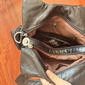 May include: Black faux leather shoulder bag with multiple compartments, including a zippered main section and interior pockets. The bag features a silver-tone charm and a brown fabric lining. The bag is open, revealing the interior.