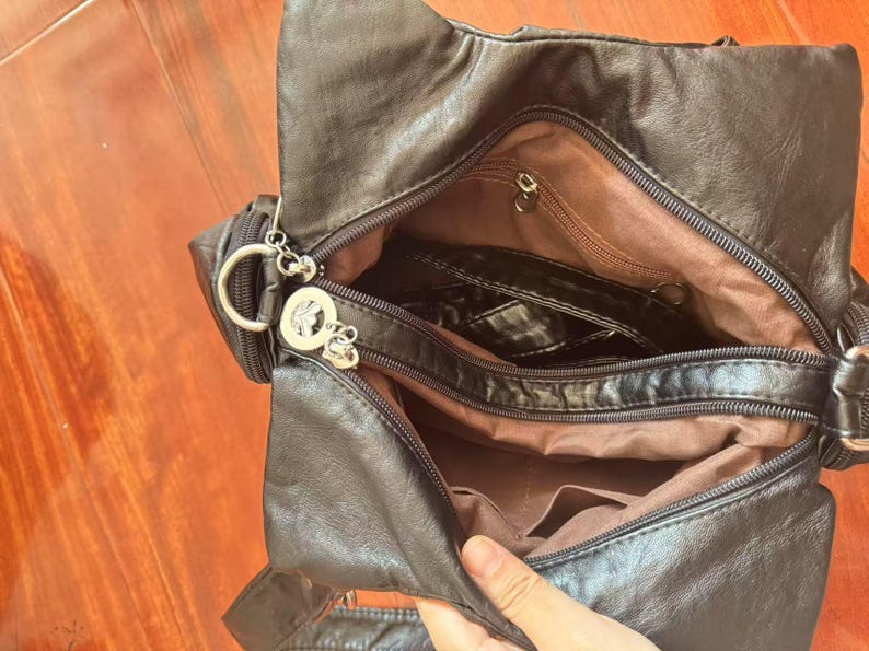 May include: Black faux leather shoulder bag with multiple compartments, including a zippered main section and interior pockets. The bag features a silver-tone charm and a brown fabric lining. The bag is open, revealing the interior.