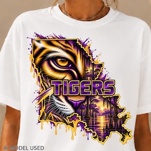 May include: A white t-shirt featuring a graphic of a tiger eye and a swamp landscape shaped like the state of Louisiana. The design includes purple and gold paint splatter effects and the word TIGERS in bold purple letters with gold outlines.