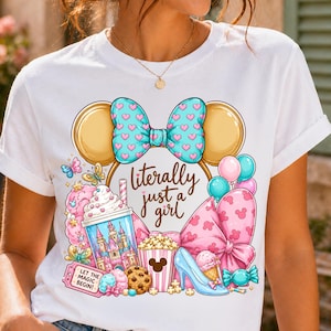Pode incluir: Camiseta branca com um design gráfico colorido. O design inclui um laço com padrões de corações, orelhas do Mickey Mouse e o texto "literally just a girl". Outros elementos incluem um castelo, sorvete e pipoca.