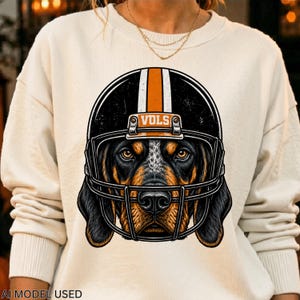 Puede incluir: Una sudadera de cuello redondo color crema con un estampado gráfico de un perro sabueso que lleva un casco de fútbol americano negro con una franja naranja y el texto VOLS. El perro tiene pelaje negro y fuego, hocico moteado y ojos marrones.
