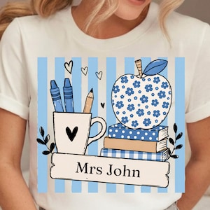 Könnte beinhalten: Weißes T-Shirt mit einem Lehrer-Themen-Design. Das Design zeigt einen blauen Blumenapfel, Bücher, eine Tasse mit Bleistiften und Buntstiften sowie den Text "Mrs John" auf einem Banner. Der Hintergrund hat blaue Streifen.