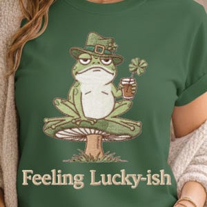 Op de afbeelding: Groen T-shirt met een geborduurde kikker die een groene hoed met een klavertje draagt en een drankje en een klavertjevier vasthoudt. De tekst "Feeling Lucky-ish" staat onderaan het shirt.
