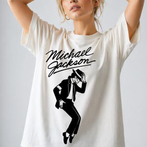 May include: Cream-colored t-shirt featuring a black graphic of Michael Jackson in a signature pose, with the name "Michael Jackson" in cursive above the image. The design is centered on the shirt.
