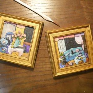 May include: Two framed miniature paintings with gold frames. One depicts a room with a blue sofa, a black cat, and a table with flowers. The other shows a room with a blue chair and a vase of flowers. A paintbrush rests nearby.