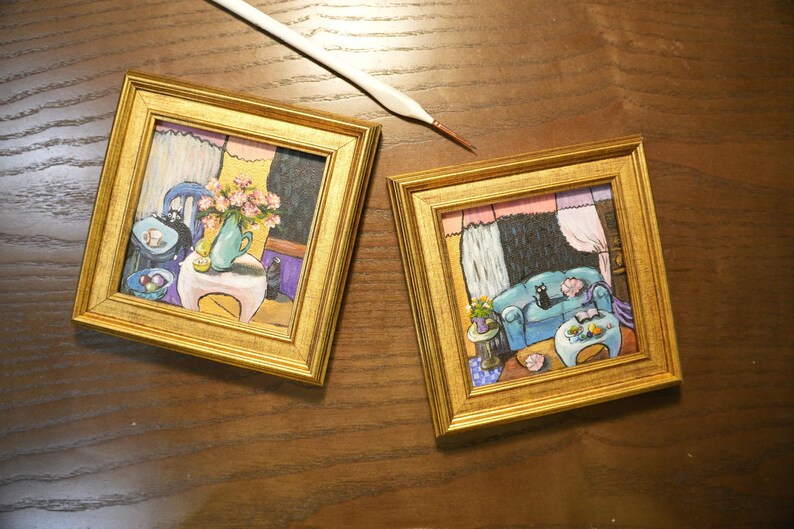 May include: Two framed miniature paintings with gold frames. One depicts a room with a blue sofa, a black cat, and a table with flowers. The other shows a room with a blue chair and a vase of flowers. A paintbrush rests nearby.