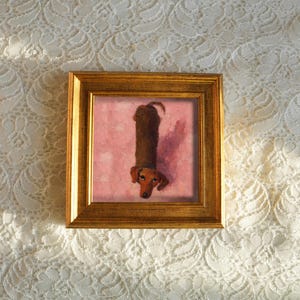 May include: A framed artwork featuring a dachshund dog. The dog is depicted in a vertical orientation against a pink background. The frame is a warm gold color, and the artwork is set against a white lace background.