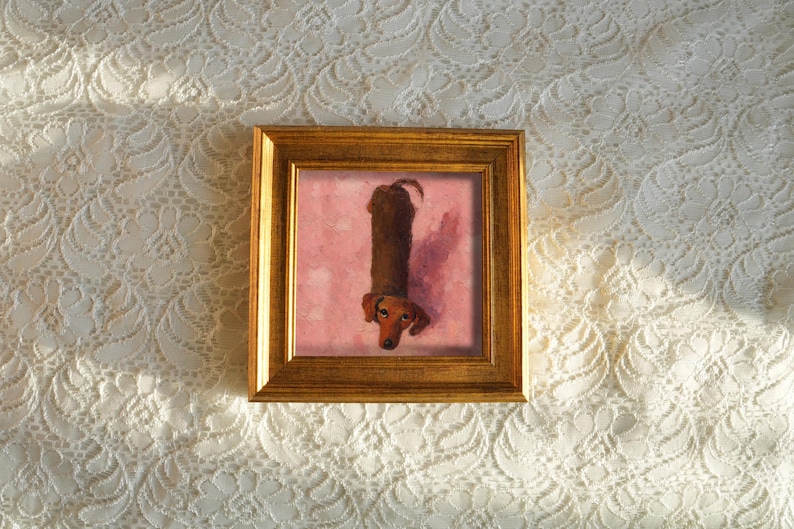 May include: A framed artwork featuring a dachshund dog. The dog is depicted in a vertical orientation against a pink background. The frame is a warm gold color, and the artwork is set against a white lace background.