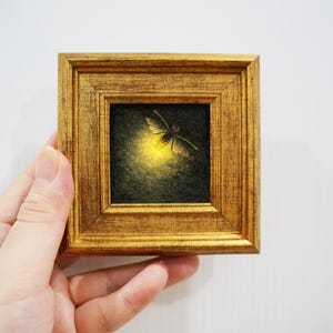 Puede incluir: Una pequeña pintura cuadrada en un marco dorado texturizado que representa una luciérnaga sobre un fondo oscuro. El insecto se muestra con el abdomen brillando en una luz amarilla intensa, creando contraste con los tonos oscuros de la escena.