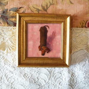 May include: A framed artwork featuring a dachshund dog. The dog is painted in shades of brown and tan, with a long body and short legs. The artwork is set against a pink background and framed in a gold-colored frame. The background is a floral pattern.