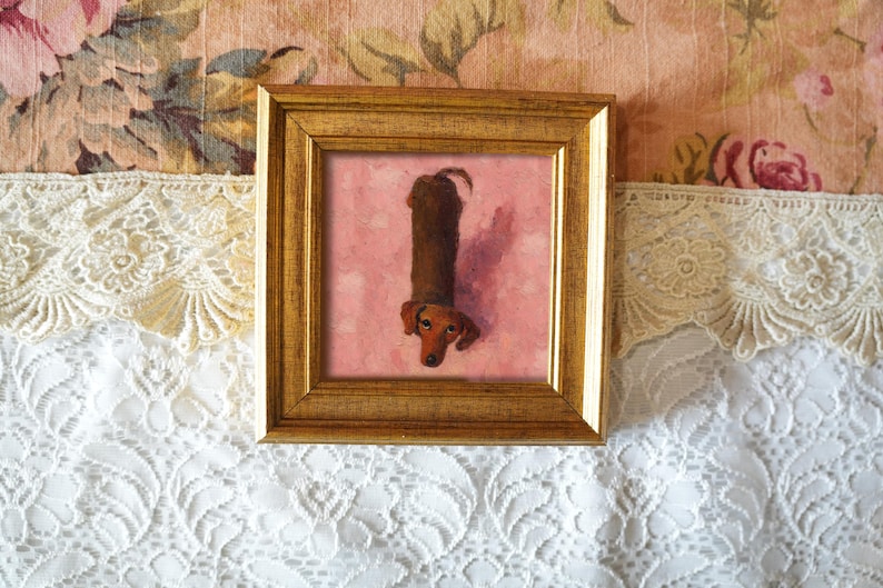 May include: A framed artwork featuring a dachshund dog. The dog is painted in shades of brown and tan, with a long body and short legs. The artwork is set against a pink background and framed in a gold-colored frame. The background is a floral pattern.