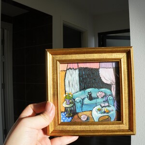 May include: A small, square painting in a gold frame depicts a cozy living room scene. A blue sofa with a black cat is the focal point. A small table with flowers and a coffee table with fruit are also visible. The artwork has a whimsical, illustrative style.