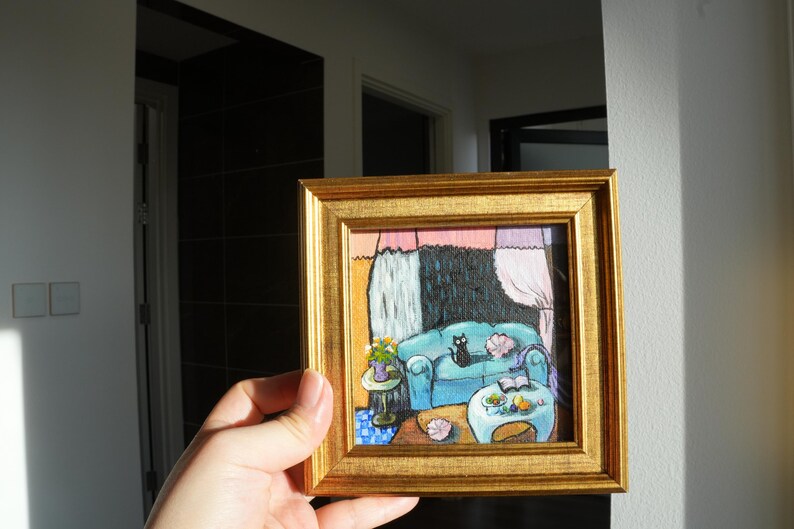 May include: A small, square painting in a gold frame depicts a cozy living room scene. A blue sofa with a black cat is the focal point. A small table with flowers and a coffee table with fruit are also visible. The artwork has a whimsical, illustrative style.