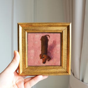 May include: A framed painting of a dachshund dog. The dog is brown with a long body and short legs, depicted against a pink background. The frame is gold-colored and square. The artwork is held up by a hand.