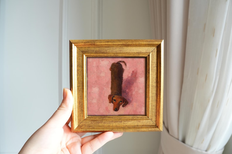 May include: A framed painting of a dachshund dog. The dog is brown with a long body and short legs, depicted against a pink background. The frame is gold-colored and square. The artwork is held up by a hand.