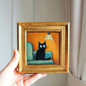 May include: A framed painting depicts a black cat with large white eyes seated on a teal couch under a lit lamp. The artwork is set against an orange background and framed in a gold-colored frame. The painting is held by a hand.