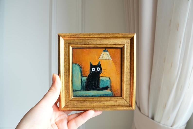 May include: A framed painting depicts a black cat with large white eyes seated on a teal couch under a lit lamp. The artwork is set against an orange background and framed in a gold-colored frame. The painting is held by a hand.