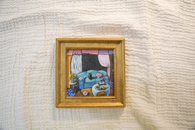 May include: A framed painting depicts a cozy interior scene. A blue sofa with a black cat is the focal point. A small table with flowers and a coffee table with fruit are also visible. The artwork is set against a backdrop of curtains and a window, all within a gold frame.