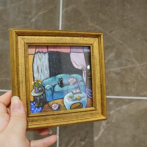 May include: A small, square painting in a gold frame depicts a cozy living room scene. A blue sofa with a black cat sits in front of a window with pink curtains. A small table holds flowers and a book. The artwork is held by a hand.