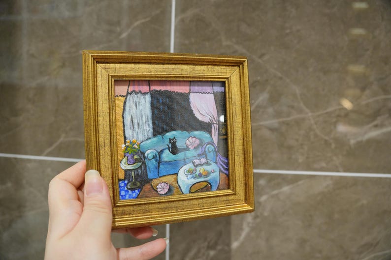 May include: A small, square painting in a gold frame depicts a cozy living room scene. A blue sofa with a black cat sits in front of a window with pink curtains. A small table holds flowers and a book. The artwork is held by a hand.