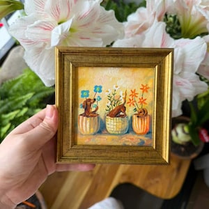 Op de afbeelding: Een klein vierkant schilderij in een gouden lijst met drie bruine teckels die in potten met patronen zitten. Elke hond is omringd door kleurrijke bloemen, waaronder blauwe, witte en oranje bloesems, tegen een zachte gele achtergrond.