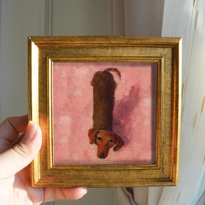 May include: A framed painting of a dachshund dog. The dog is depicted in a vertical orientation, with a brown body and head, and a pink background. The frame is gold-colored.