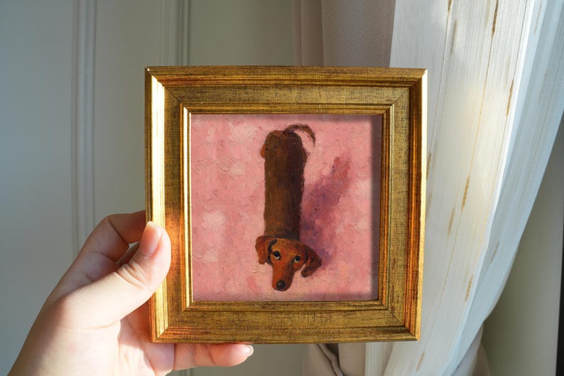 May include: A framed painting of a dachshund dog. The dog is depicted in a vertical orientation, with a brown body and head, and a pink background. The frame is gold-colored.